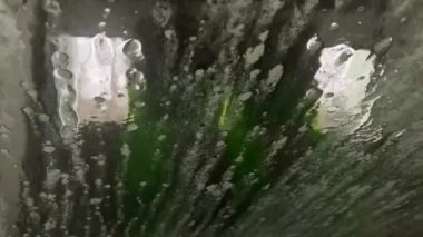 İçerideki su akışı arabanın ön camındaki köpüğü ve kiri silip süpürüyor. Otomatik oto yıkama işlemi. Araba camında sabun köpükleri. Beyaz sabun köpüğü araba camında hareket eder..