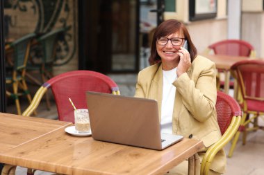 Olgun bir ofis çalışanı, elinde bir fincan kahve ve dizüstü bilgisayarla terasta otururken akıllı telefonuyla yaklaşan iş seyahatinden bahseder.