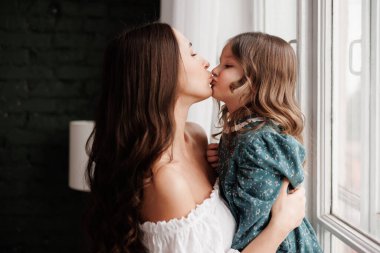 Anneler Günü. Mutlu genç anne, sevimli kızıyla pencerenin önünde kucaklaşıyor. Anneyle çocuk arasındaki güven, destek ve sevgi. Sevgi dolu aile tatlı anların tadını çıkarır. Çocuk günü