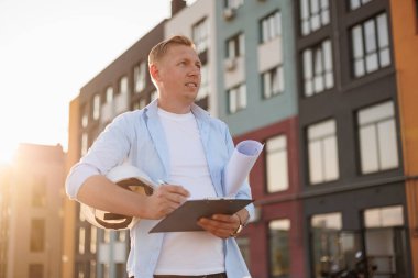 Erkek mimar ya da müteahhit modern konut binasının hazır yeni projesinin güvenliğini inceliyor ve inceliyor. Profesyonel inşaat mühendisi planları tutar ve dışarıdaki panolarda not tutar.