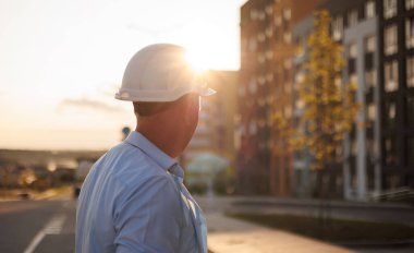 Erkek mimarın arka görünüşü, koruyucu kask takan müteahhit, modern konut inşaatının hazır yeni projesinin güvenliğini inceliyor. İnşaat mühendisi, işçi ustabaşı, güneş ışınları.