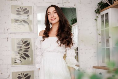 Kadınlar Günü. 20-25 yaşlarında, uzun kıvırcık saçlı, çapraz elli romantik ve çekici bir genç kadın, arka plandaki modern moda ev mutfağına bakıyor.