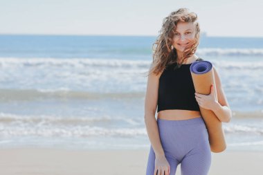 Genç kadın yoga minderini tutuyor, kameraya bakıyor ve gülümsüyor. Bayan dışarıda antrenman yapmaya hazırlanıyor. Deniz kıyısında spor ve spor. Sağlıklı yaşam tarzı, sabah egzersizleri, kadın sağlığı ve gücü, sağlık hizmetleri..
