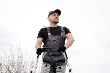 Koruyucu giysili, eldivenli, tulumlu profesyonel erkek bahçıvanın alt görünüşü, dışarıdaki çimleri biçmek için kablosuz benzin çim biçme makinesini tutuyor. Mevsimlik peyzaj tasarımı