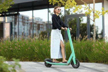 Mutlu stil sahibi kadın, güneşli yaz gününde iş bölgesinde elektrikli scooter ya da e-scooter kullanıyor. Modern çevre dostu yaşam tarzı kavramı ve sürdürülebilirlik kentsel yaşam, motorlu araç