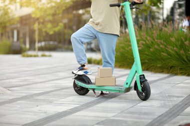 Park halindeki elektrikli scooter 'ın yanında sıradan kıyafetler giymiş, iş yerindeki yeşil caddede karton kutular taşıyan tanınmamış bir adam. Sürdürülebilirlik kavramı, dağıtım ve çevre dostu modern yaşam tarzı