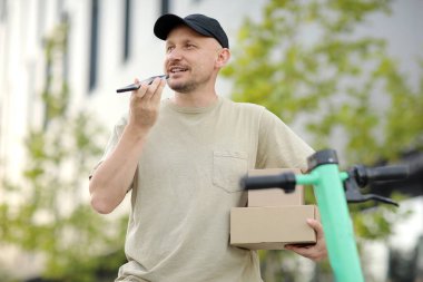 Telefon kullanan genç teslimatçının portresi açık alanda park halindeki scooter 'ın yanında karton kutular tutuyor. Sürdürülebilirlik kavramı, dağıtım ve çevre dostu modern yaşam tarzı