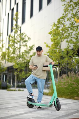 Telefon kullanan genç kurye, park halindeki elektrikli scooter 'ın yakınındaki müşteriyi arıyor iş alanındaki caddede karton kutular teslim ediyor. Sürdürülebilirlik kavramı, dağıtım ve çevre dostu modern yaşam tarzı