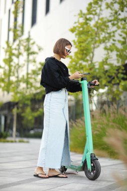 Akıllı telefonu olan şık bir genç kadın, elektrikli scooter kiralama için QR kodunu tarıyor. Sokaktaki limansız ulaşımı açmak için telefondaki uygulamayı kullanan bir kız. Kentsel tarzda ekolojik araç kullanmak. Modern teknoloji