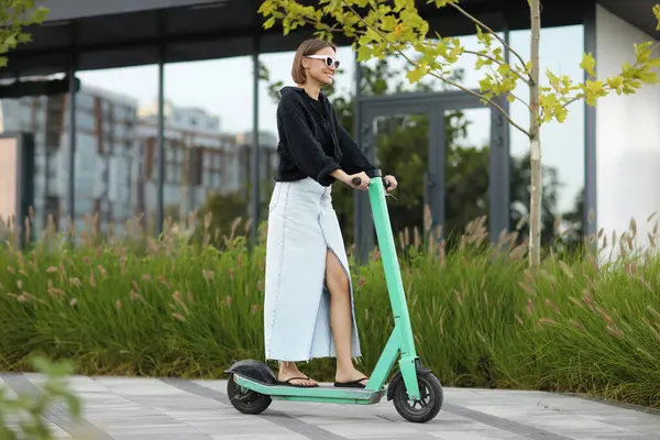 Mutlu stil sahibi kadın, güneşli yaz gününde iş bölgesinde elektrikli scooter ya da e-scooter kullanıyor. Modern çevre dostu yaşam tarzı kavramı ve sürdürülebilirlik kentsel yaşam, motorlu araç