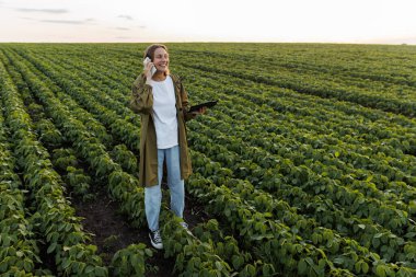 Modern tarım sektörü. Dijital tabletli mutlu kadın çiftçi, tarladaki soya fasulyesi bitkilerini inceledikten sonra akıllı telefonla konuşuyor. Agronomist hasatların büyümesini kontrol eder. Akıllı tarım teknolojisi.