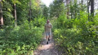 Yeşil orman yolunda kablosuz kulaklıklarla koşan ve yazın müzik dinleyen yetişkin bir adamın ön görüntüsü. Aktif yaşam tarzı, sağlıklı alışkanlıklar ve motivasyon konsepti. Doğada egzersiz..