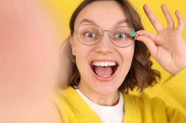 Gözlüklü mutlu genç bir kadının portresi, selfie çekiyor ve sarı arka planda kameraya geniş bir şekilde gülümsüyor. Terfi kavramı, kişisel markalaşma, dijital pazarlama, çevrimiçi eğitim içeriği