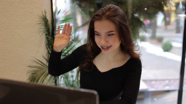 Genç kadın kafede dizüstü bilgisayar kullanıyor. Video çağrısı ve el sallamalar, internette konuşmalar, gülümsemeler. Modern teknoloji, gençlik yaşam tarzı, Z jenerasyonu. Uzaktan çalışma, e-öğrenme, çevrimiçi uzaktan çalışma.