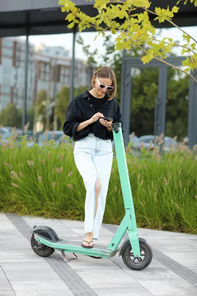 Şık bir kadın akıllı telefondan iletişim kuruyor. Güneşli yaz günlerinde iş bölgesinde elektrikli scooter ya da e-scooterın yanında duruyor. Modern çevre dostu yaşam tarzı ve sürdürülebilirlik kentsel yaşam kavramı
