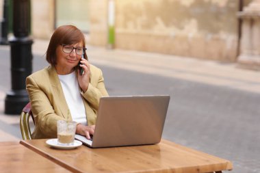 Olgun bir ofis çalışanı, elinde bir fincan kahve ve dizüstü bilgisayarla terasta otururken akıllı telefonuyla yaklaşan iş seyahatinden bahseder.