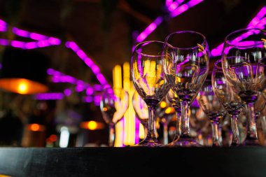Boş, berrak şarap kadehleri gece kulübündeki bar tezgahında duruyor. Arka planda bulanık mor neon ve sarı ışıklar var. Temiz kokteyl kadehleri kutlama için hazır..