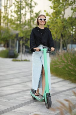 Mutlu stil sahibi kadın, güneşli yaz gününde iş bölgesinde elektrikli scooter ya da e-scooter kullanıyor. Modern çevre dostu yaşam tarzı kavramı ve sürdürülebilirlik kentsel yaşam, motorlu araç