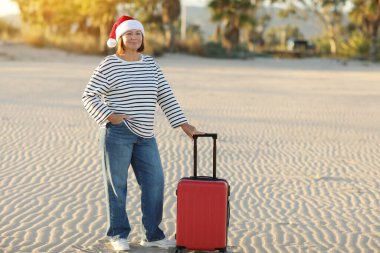 Noel Baba şapkalı, çizgili tişörtlü, 50-55 yaş arası mutlu bir yetişkin kadın kum sahili kenarında kırmızı bavul tutuyor. Palmiyeli yeni yıl tatilini tropikal tatil beldesinde yalnız kutluyor..