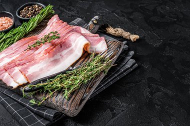 Raw smoked bacon slices with rosemary and thyme on wooden board. Black background. Top view. Copy space.