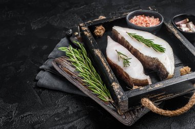 Raw halibut fish steak in wooden tray with herbs. Black background. Top view. Copy space.