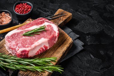 Raw Rib-eye Steak, beef marbled meat on wooden board with rosemary. Black background. Top view. Copy space.