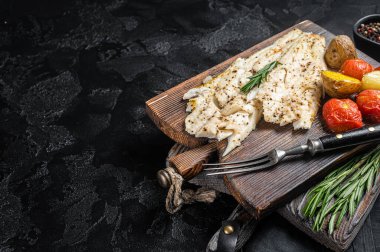 Baked Haddock fish fillet on wooden board with tomato and potato. Black background. Top view. Copy space.
