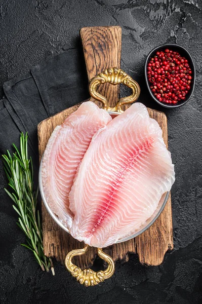 Bitkilerle dolu bir tavada çiğ tilapia balığı filetosu pişirmeye hazırım. Siyah arka plan. Üst görünüm.