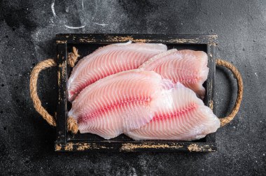 Fresh tilapia white fish fillet in a wooden tray. Black background. Top view.