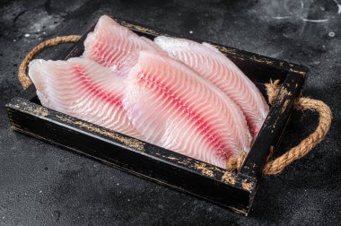 Fresh tilapia white fish fillet in a wooden tray. Black background. Top view.