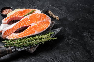 Uncooked Raw Salmon Fish Steaks on butcher board with herbs. Black background. Top view. Copy space.