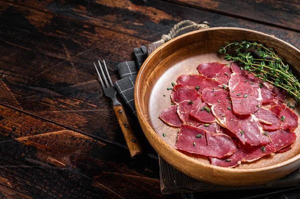 Pastrami slices, dried beef meat with herbs in wooden plate. Wooden background. Top view. Copy space.