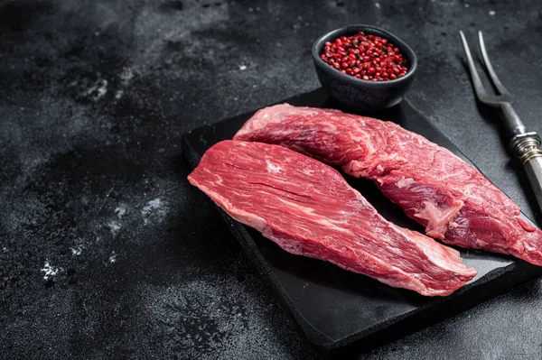 Bavette raw beef meat steak or Sirloin flap on marble board. Black background. Top view. Copy space.