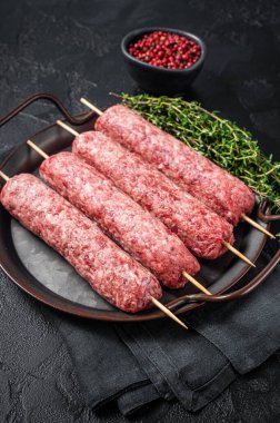 Raw Turkish Urfa kebab skewers in steel tray with herbs. Black background. Top view.