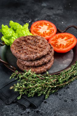 Tasty grilled burger beef patty with tomato, spices and lettuce in kitchen tray. Black background. Top view.