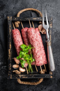 Fresh raw beef meat kebabs sausages on skewers in wooden tray. Black background. Top view.