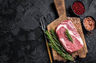 Raw Rib-eye Steak, beef marbled meat on wooden board with rosemary. Black background. Top view. Copy space.