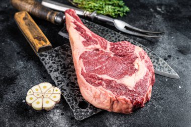 Dry aged raw tomahawk or cowboy beef steak on a black table with knife, cleaver, spice and herbs. Black background. Top view.