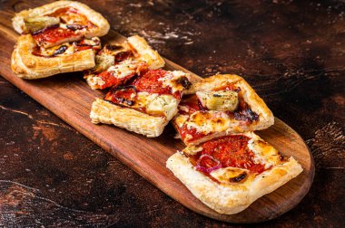 Baked Puff pastry tart pizza with artichoke, mozzarella, tomatoes and cheese sliced on a wooden board. Dark background. Top view.