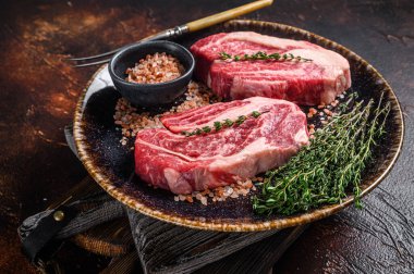 Chuck Eye Roll bifteği, kekik ve otlarla dolu bir tabakta çiğ et. Karanlık arka plan. Üst görünüm.