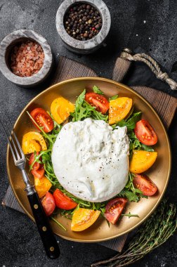 Burrata cheese with cherry tomatoes and fresh arugula in a plate. Black background. Top view.