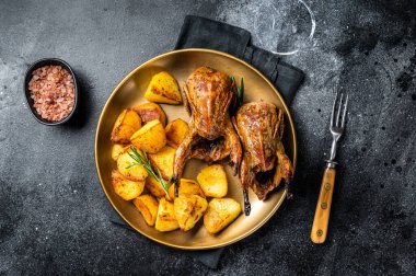 Delicious roasted quails with herbs in a plate with fried potato. Black background. Top view.