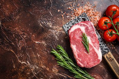 Uncooked raw Rib eye Steak, ribeye beef meat on butcher cleaver with herbs. Dark background. Top view. Copy space.