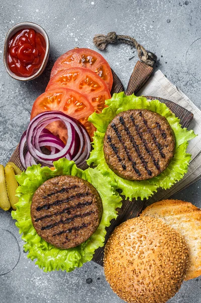 Plant based meatless burgers with vegan grilled pattie, tomato and onion on a wooden serving board. Gray background. Top view.