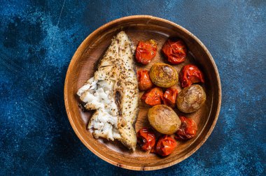 Baked halibut fish with roasted tomato and potato in wooden plate. Blue background. Top view. Copy space.