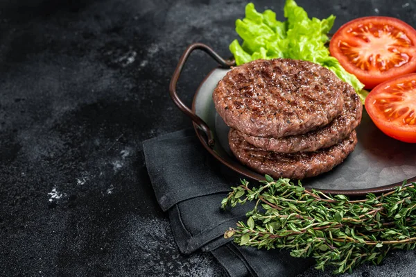 Tasty grilled burger beef patty with tomato, spices and lettuce in kitchen tray. Black background. Top view. Copy space.