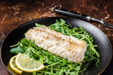 Grilled cod fish fillet served with green salad in a plate. Dark background. Top view.