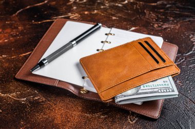 Wooden desk table with blank notebook, pen, wallet with money and credit cards. Brown background. Top view. Copy space.