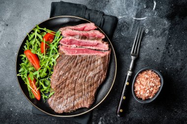 Grilled medium rare flank beef steak with salad in a plate. Black background. Top view.