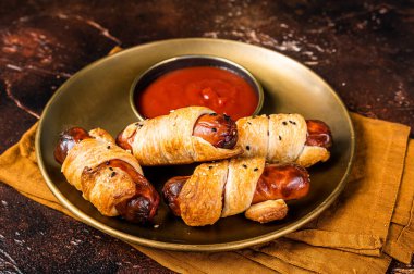 Rolled hot dog sausages baked in puff pastry with ketchup. Dark background. Top view.